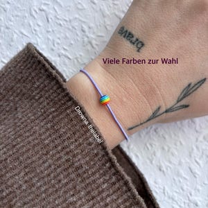Könnte beinhalten: Ein lila Armband mit einer Regenbogenperle. Das Armband befindet sich an einem Handgelenk mit einem "brave" Tattoo. Der Text "Viele Farben zur Wahl" ist ebenfalls sichtbar. Der Markenname "Drowna.thelabel" ist auf dem Bild aufgedruckt.