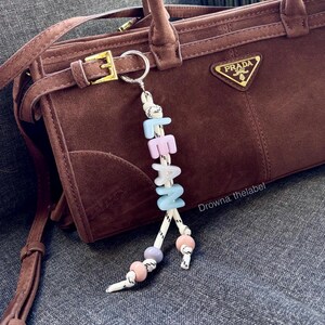 Könnte beinhalten: Eine braune Wildlederhandtasche mit goldenem Prada-Logo-Schild und Schnallenriemen, ergänzt durch einen weißen Kordel-Schlüsselanhänger mit pastellblauen und rosa Buchstabenperlen, die LEAN buchstabieren, und runden Pastellperlen an den Enden.