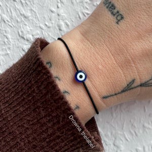 Könnte beinhalten: Ein schwarzes Armband aus Schnur mit einem blauen und weißen bösen Blick-Anhänger.