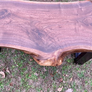 May include: A large, live edge walnut wood bench with a natural, dark brown finish. The bench has a curved top and is supported by two black metal legs.