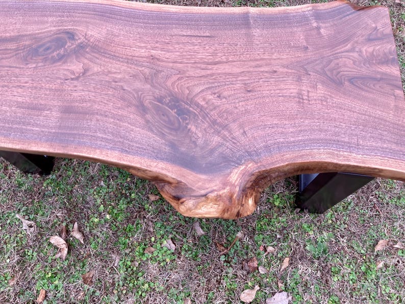 May include: A large, live edge walnut wood bench with a natural, dark brown finish. The bench has a curved top and is supported by two black metal legs.