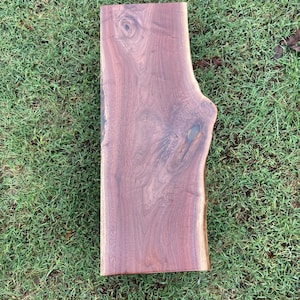 May include: A large, rectangular slab of wood with a natural, live edge. The wood has a rich, dark brown color and a smooth, polished finish. The grain of the wood is visible and adds to the natural beauty of the piece.