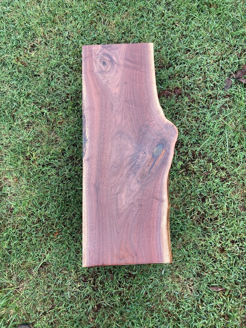 May include: A large, rectangular slab of wood with a natural, live edge. The wood has a rich, dark brown color and a smooth, polished finish. The grain of the wood is visible and adds to the natural beauty of the piece.
