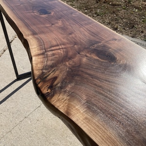 May include: A live edge walnut wood console table with a dark brown finish. The table has a natural, irregular edge and is supported by a black metal frame. The wood grain is visible, adding to the rustic aesthetic.