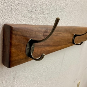 May include: A wooden wall-mounted coat rack with three metal hooks. The rack is made of dark brown wood and has a natural finish. The hooks are a brushed nickel color.