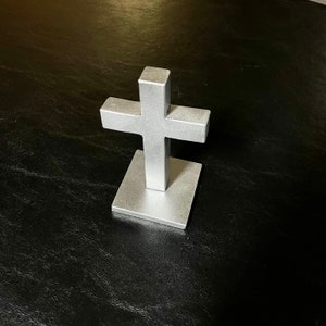 May include: A silver metal cross with a square base. The cross is standing on a black surface.