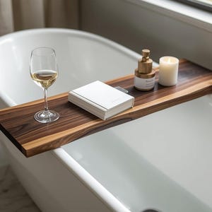 May include: A wooden bathtub tray with a glass of white wine, a white book, a candle, and a bottle of perfume. The tray is made of dark brown wood and is placed across a white bathtub. The items on the tray create a relaxing spa-like atmosphere.