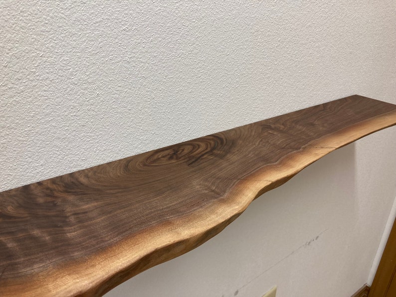 May include: A live edge wooden shelf featuring a dark brown walnut grain with visible swirling patterns and a natural, irregular bark-free edge. The horizontal board is mounted against a textured white wall, showcasing the wood's organic texture.