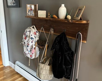 May include: A rustic wooden shelf with metal hooks, holding a floral backpack, purses, a black jacket, and a dog leash. The shelf also displays decorative items, including a vase, framed photos, and a sign that says "HIPPITY HOPPITY".