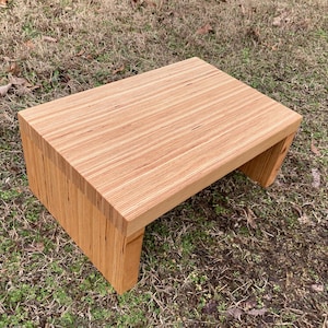 May include: A wooden step stool with a natural wood finish. The stool is made of plywood and has a rectangular top.