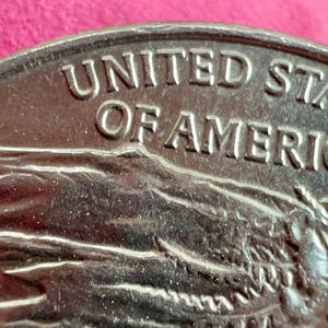May include: Close-up of a silver coin with the words "UNITED STATES OF AMERICA" embossed on the top edge. The coin's surface shows a detailed relief of a landscape and other design elements.
