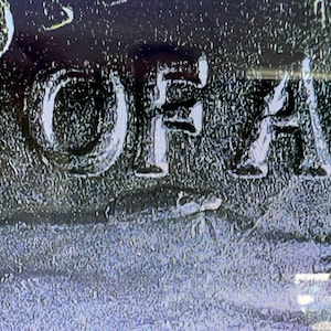 May include: Close-up of a textured, dark surface with the word "OF" and part of another word in white, raised lettering. The image has a grainy, almost metallic appearance, with subtle variations in color.