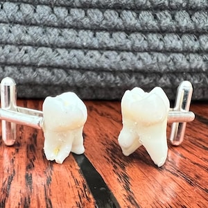 May include: A pair of silver cufflinks with white tooth-shaped charms. The cufflinks are on a wooden surface.