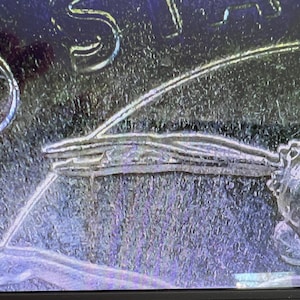 May include: Close-up of a silver coin with intricate details. The coin features a detailed design with curved lines and a textured surface. The image highlights the coin's craftsmanship and the fine lines of the design.