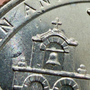 May include: Close-up of a silver coin featuring a detailed depiction of a building with arches, a bell, and a cross. The word "ANTON" is visible at the top of the coin.