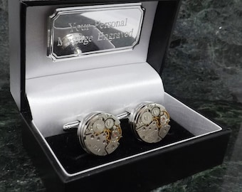 Puede incluir: Un par de gemelos de plata con un diseño de movimiento de reloj, presentados en una caja negra con una placa de plata que dice "Your Personal Message Engraved".