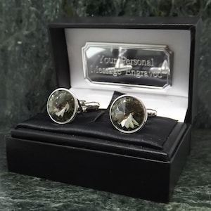 Puede incluir: Un par de gemelos de plata con piedras de cristal transparentes en una caja de regalo negra. La caja tiene una placa de plata con el texto "Your Personal Message Engraved".