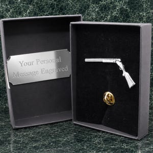 Peut inclure: Une épinglette en forme de fusil de couleur argentée présentée dans une boîte grise. La boîte comporte une plaque argentée avec le texte gravé Your Personal Message Engraved. L'épinglette comprend une attache papillon dorée.
