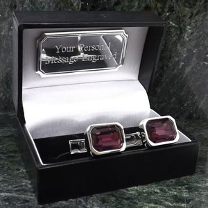 May include: A pair of silver cufflinks with a dark purple rectangular gemstone in each. The cufflinks are in a black box with a silver plate that reads "Your Personal Message Engraved".