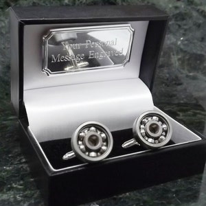 Puede incluir: Gemelos de plata en una caja de regalo negra. Los gemelos tienen forma de rodamientos de bolas. La caja tiene una placa de plata que dice "Su mensaje personal grabado".