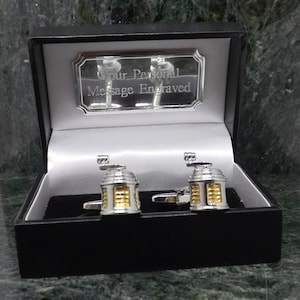 Może przedstawiać: Czarne pudełko prezentowe z srebrną tabliczką z napisem "Your Personal Message Engraved". Pudełko jest otwarte i zawiera dwa srebrne spinki do mankietów z złotymi akcentami. Spinki do mankietów mają kształt małych budynków.