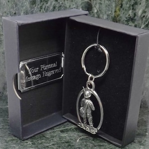 Może przedstawiać: Srebrny brelok do kluczy z detalicznym wizerunkiem osoby trzymającej torbę i kij. Brelok znajduje się w czarnym pudełku z srebrną tabliczką z napisem "Twój osobisty grawerowany komunikat".