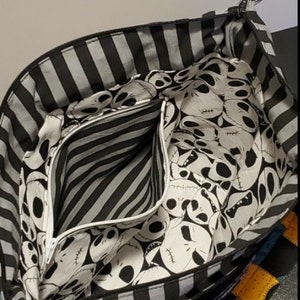 May include: Black and white striped tote bag with a zippered pocket. The lining of the bag features a repeating pattern of Jack Skellington's face from the Nightmare Before Christmas.