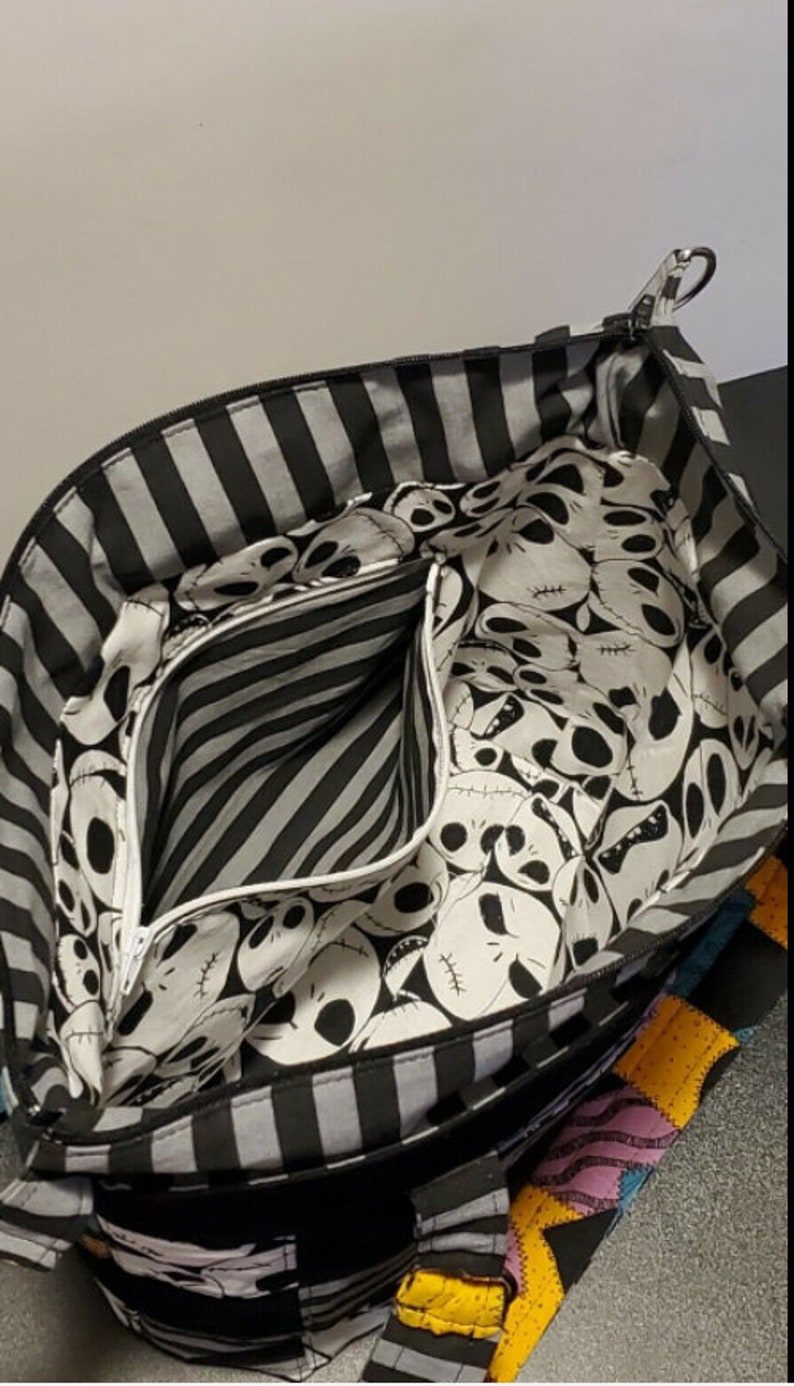 May include: Black and white striped tote bag with a zippered pocket. The lining of the bag features a repeating pattern of Jack Skellington's face from the Nightmare Before Christmas.
