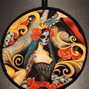 May include: Round black pot holder with a colorful design featuring a woman with a sugar skull face, roses, and skulls. The background is black with gold swirls.