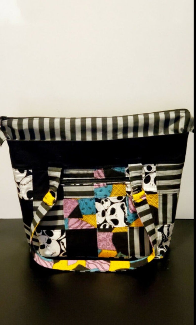 May include: A black and white striped tote bag with a patchwork design featuring characters from the Nightmare Before Christmas. The bag has a zipper closure and two yellow straps.