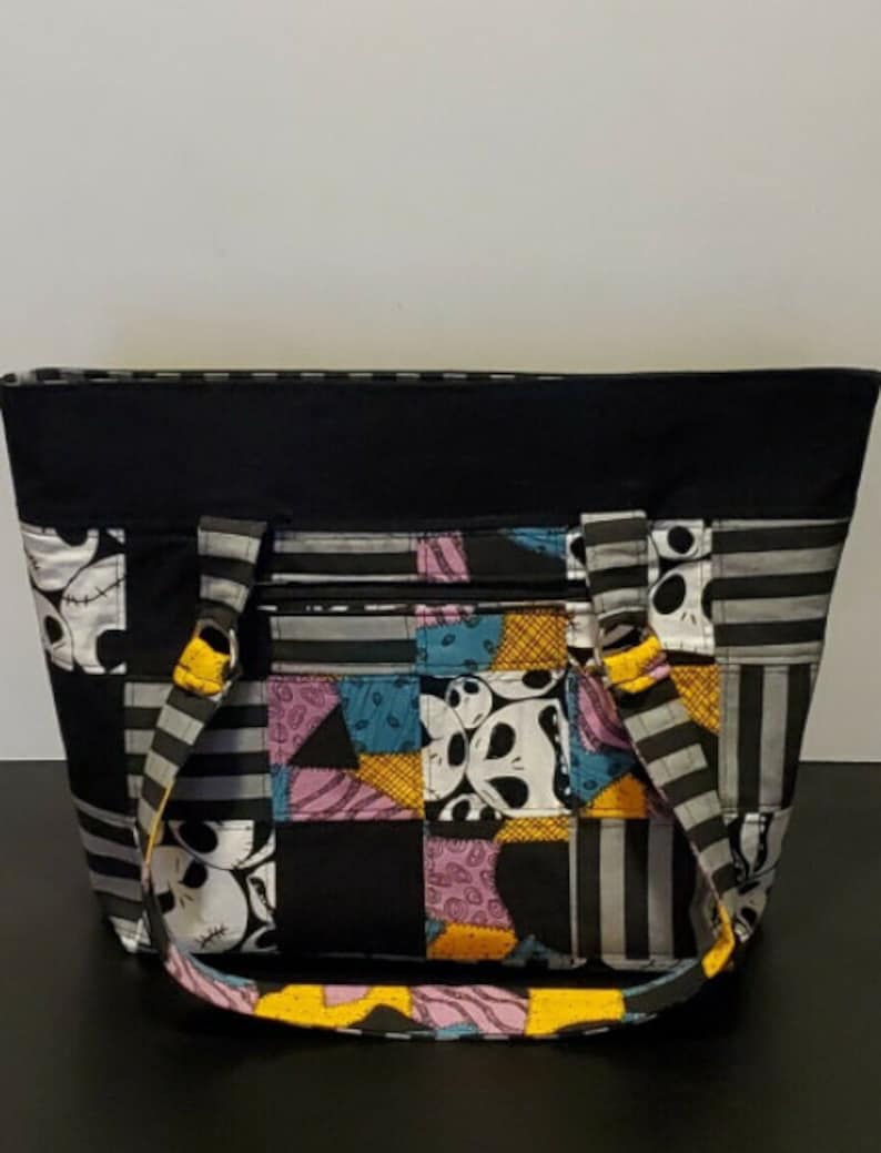 May include: Black tote bag with a patchwork design featuring a character from the Nightmare Before Christmas. The bag has yellow straps and a black zipper.