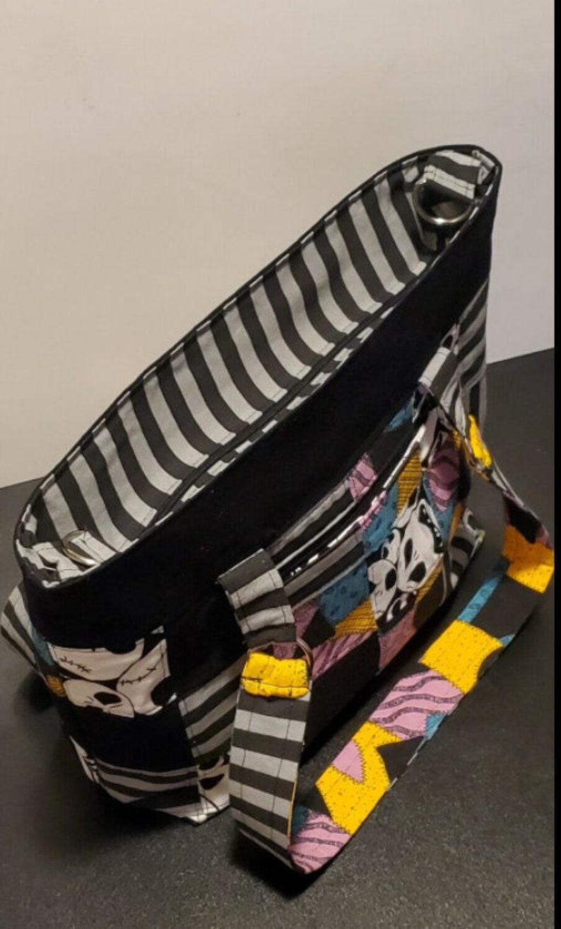May include: Black and white striped tote bag with a patchwork design featuring characters from The Nightmare Before Christmas. The bag has two black handles and a zipper closure.