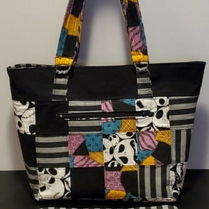 May include: A black tote bag with a patchwork design featuring a character from the Nightmare Before Christmas. The bag has a zippered pocket and a striped shoulder strap.