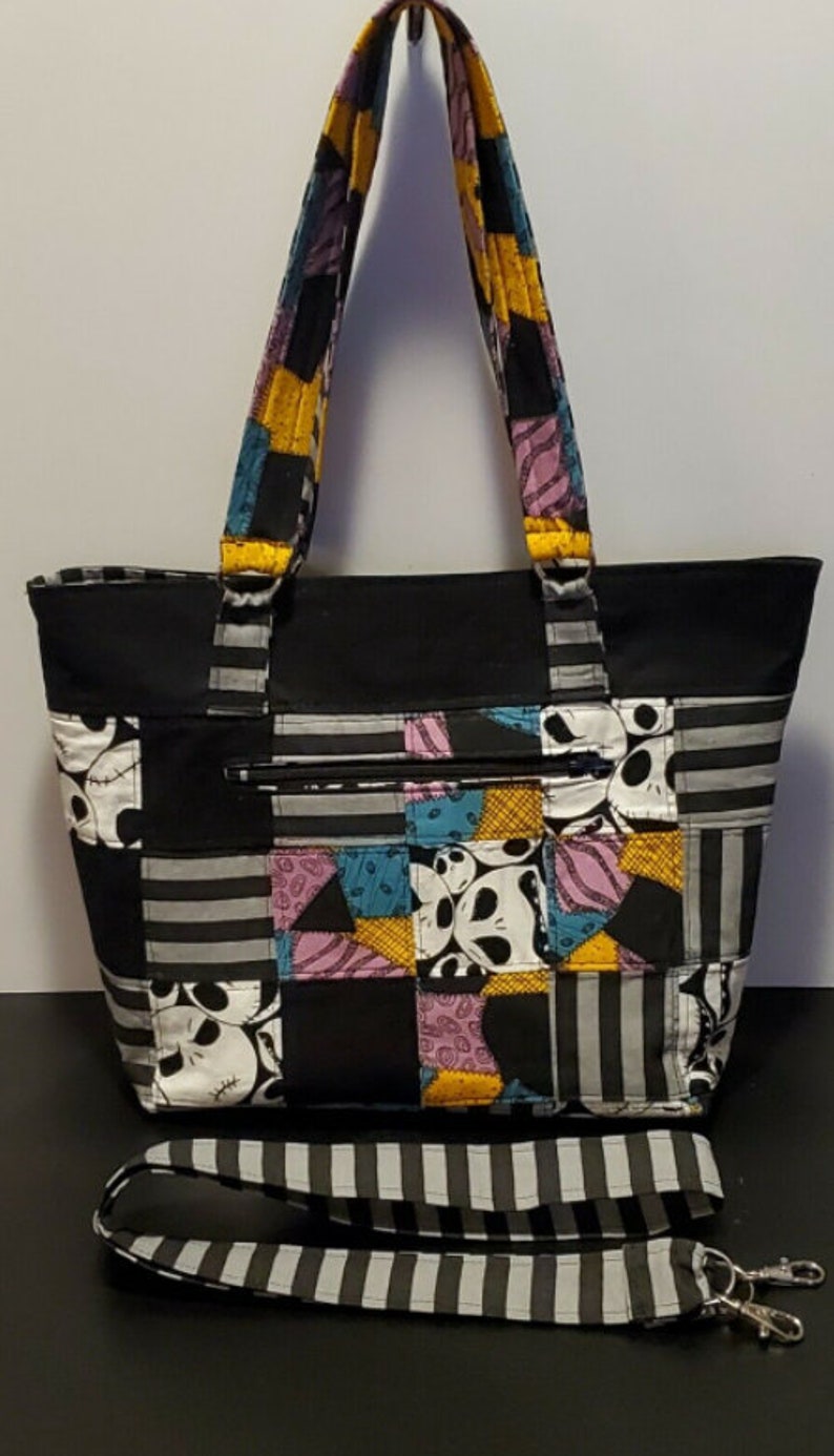 May include: A black tote bag with a patchwork design featuring a character from the Nightmare Before Christmas. The bag has a zippered pocket and a striped shoulder strap.