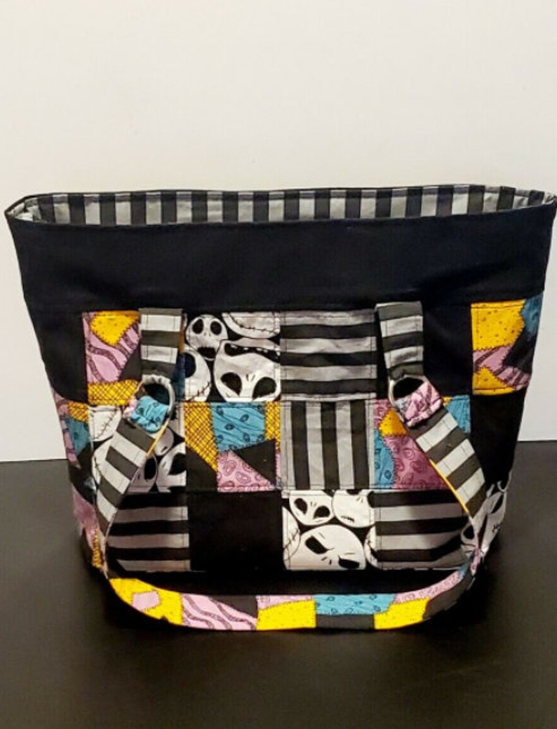 May include: A black patchwork tote bag with a striped lining. The bag features a patchwork design of black, white, gray, yellow, pink, and blue fabric with a repeated pattern of Jack Skellington's face from the Nightmare Before Christmas.