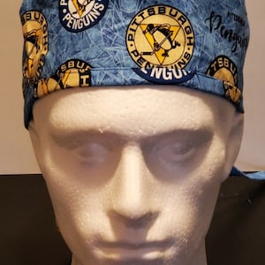 May include: Blue and yellow surgical scrub hat with a repeating pattern of the Pittsburgh Penguins logo. The logo features a penguin holding a hockey stick. The text "Pittsburgh Penguins" is written in a curved arc around the logo.