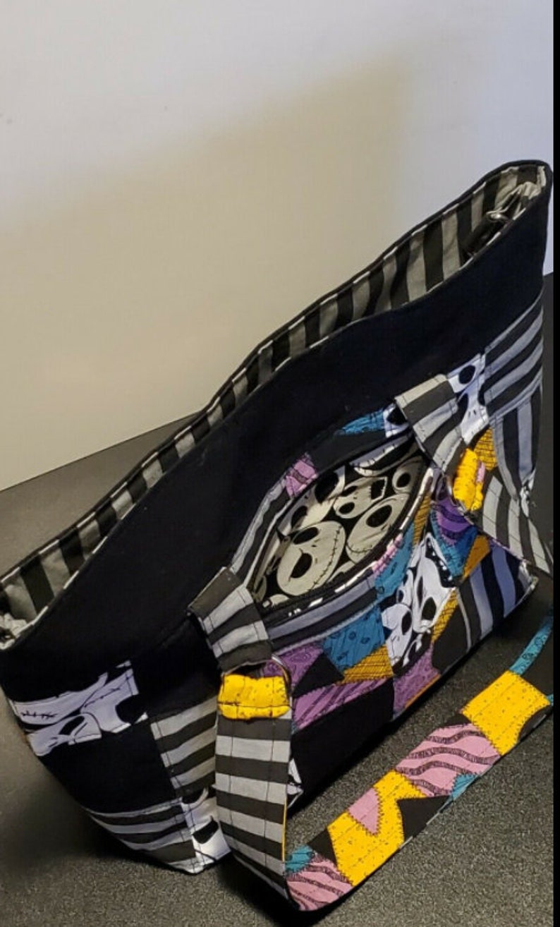 May include: Black and white striped tote bag with a patchwork design featuring characters from The Nightmare Before Christmas. The bag has a black lining and a yellow and black striped strap.