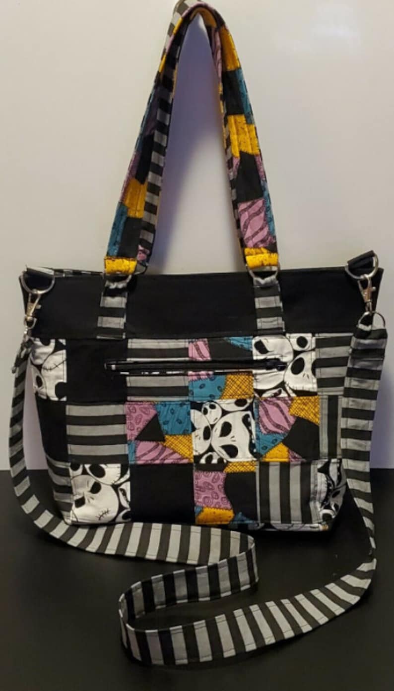 May include: A black and white striped tote bag with a patchwork design featuring Jack Skellington's face. The bag has a zippered pocket and a detachable shoulder strap.