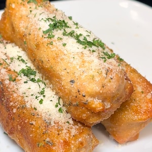 May include: Three golden brown fried spring rolls topped with grated parmesan cheese and chopped parsley on a white plate.