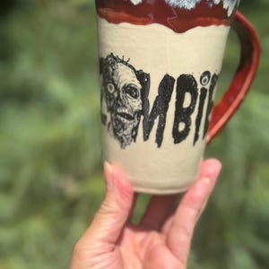 May include: A ceramic mug with a zombie graphic and the word "ZOMBIE" printed in black. The mug has a red handle and rim, with a cream-colored body. The mug is held in a person's hand.