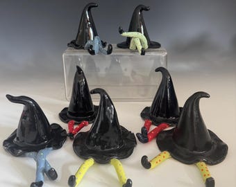 May include: A collection of seven ceramic witch hat figurines, each featuring a glossy black pointed hat with a wide brim and a pair of dangling legs. The legs are painted in various colors including light blue, yellow, and red, all with polka dots.