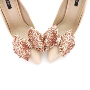May include: A pair of nude high-heeled shoes with a rose gold glitter bow on each shoe.