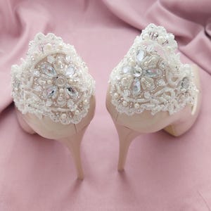 May include: A pair of beige high-heeled shoes with detailed white lace and bead embellishments. The lace design has a floral pattern with clear and pearl-like beads. The heels are slender and the background is a soft pink.