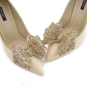 Puede incluir: Un par de zapatos de tac&oacute;n alto beige con punta. Cada zapato presenta un gran lazo dorado brillante en la puntera. Los zapatos tienen un acabado brillante y est&aacute;n sobre un fondo blanco.