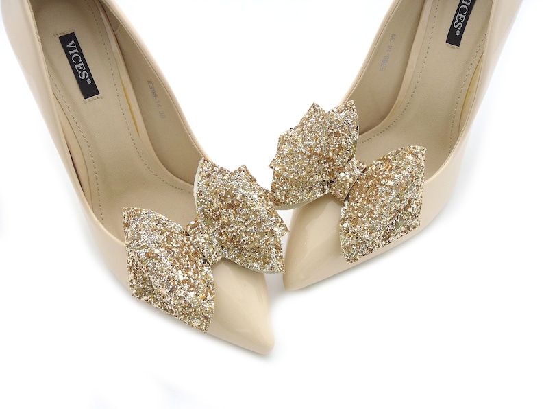 Puede incluir: Un par de zapatos de tac&oacute;n alto beige con punta. Cada zapato presenta un gran lazo dorado brillante en la puntera. Los zapatos tienen un acabado brillante y est&aacute;n sobre un fondo blanco.