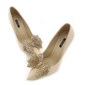 Puede incluir: Un par de zapatos de tac&oacute;n alto beige con punta. Cada zapato presenta un lazo dorado brillante en la parte delantera. La marca "VICES" est&aacute; impresa en la plantilla.