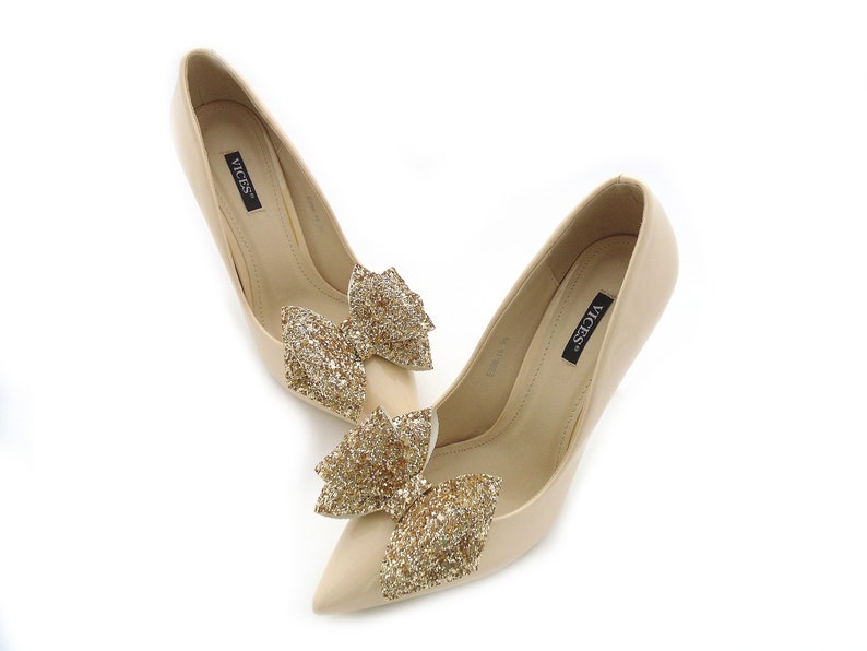 Puede incluir: Un par de zapatos de tac&oacute;n alto beige con punta. Cada zapato presenta un lazo dorado brillante en la parte delantera. La marca "VICES" est&aacute; impresa en la plantilla.