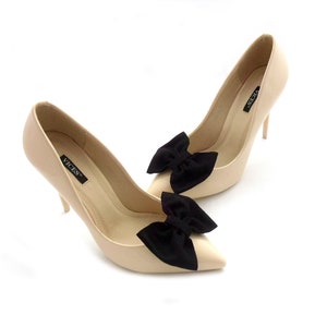 K&ouml;nnte beinhalten: Ein Paar beigefarbene Pumps mit hohen Abs&auml;tzen und schwarzen Schleifen an den Zehen. Die Schuhe sind mit "VICES" gekennzeichnet.