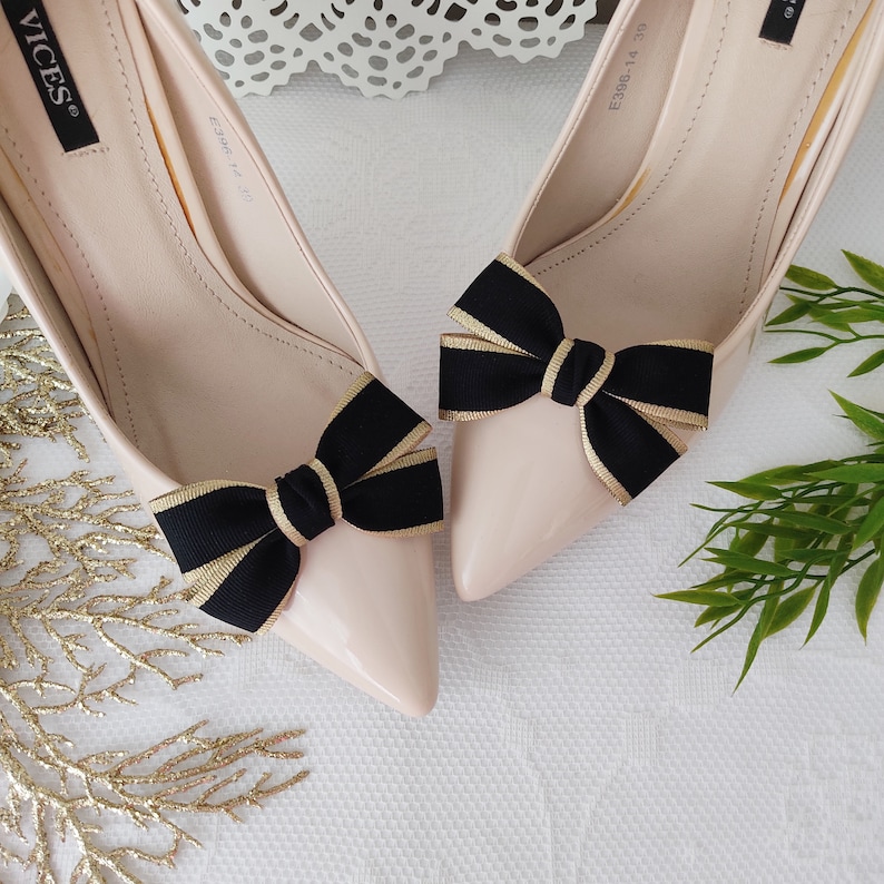May include: A pair of light pink high-heeled shoes with black and gold bows on the toes. The shoes are made of patent leather and have a pointed toe.