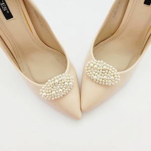 May include: A pair of beige high-heeled shoes with pearl embellishments on the toes. The shoes have a pointed toe and a stiletto heel.